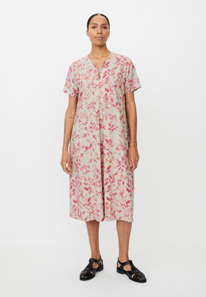 Kobieta stojąca w beżowej midi sukience w różowy kwiatowy wzór, z krótkimi rękawami i czarnymi sandałami zakrywającymi palce, na białym tle.