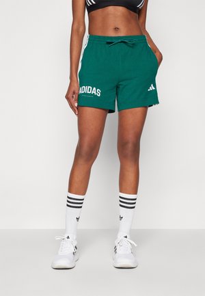 Sportowiec w zielonych spodenkach Adidas, czarnym krótkim topie, białych skarpetkach Adidas z czarnymi paskami oraz białych butach sportowych Adidas stoi na białym tle.