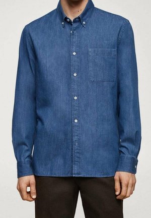 Mand iført en langærmet blå denim-tryk-knap-skjorte med brystlomme og mørke bukser, står mod en ensfarvet baggrund.