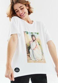 Bílý bavlněný tričko s velkým potiskem znázorňujícím sedící ženu v klasickém oblečení a černou značkovou etikou na lemu.