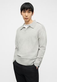 Νέος άνδρας με μαύρα μαλλιά που φορά ανοιχτό γκρι πουλόβερ με γιακά πάνω από λευκό πουκάμισο και μαύρο παντελόνι, όρθιος με τα χέρια στις τσέπες.