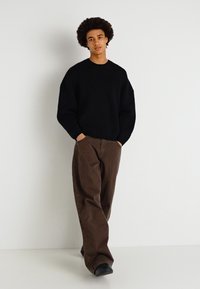 Pull en tricot noir à coupe ample, col rond ; associé à un pantalon large marron ; le modèle se tient devant un arrière-plan uni.