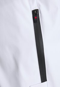 Tejido blanco con una cremallera negra y un pequeño logotipo de hoja de arce roja en el tirador de la cremallera. Textura suave con costuras contrastantes.
