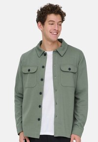 Junger Mann mit lockigem Haar, der lächelt, trägt eine ungeknöpfte olivgrüne Jacke über einem weißen T-Shirt und steht vor einem schlichten hellen Hintergrund.