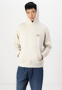 Beige hoodie met een voorste kangaroozak, ribgebreide manchetten en een logo op de linkerborst. Gecombineerd met blauwe denim jeans.