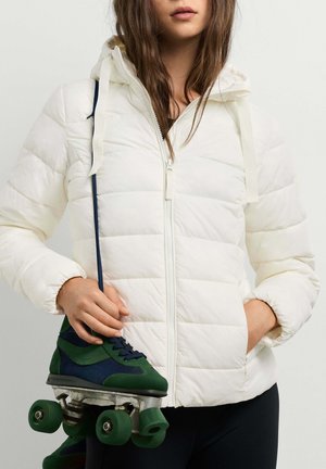 Frau, die eine weiße Steppjacke trägt, hält grüne und blaue Rollschuhe an den Schnürsenkeln mit einer Hand, die andere Hand in der Jackentasche.