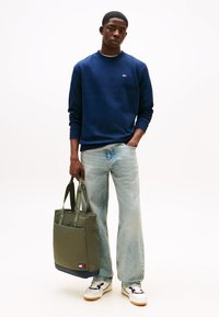 Fiatal férfi sötétkék pulóverben és világoskék farmerben, kezében oliva zöld tote táska márkás pántokkal és fehér sportcipővel.