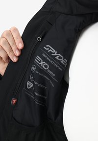 Mano sosteniendo una chaqueta Spyder negra abierta, mostrando la etiqueta interior con características de rendimiento impermeable, elástico y de aislamiento térmico.