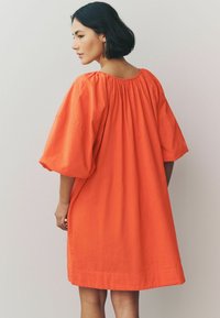 Pomarańczowa lniana sukienka o luźnym kroju, z krótkimi, bufiastymi rękawami i marszczonym dekoltem, sięgająca powyżej kolan.