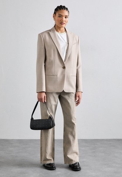 Beige maßgeschneiderter Blazer mit einem Knopf, kombiniert mit hellgrauen Hosen. Schwarze Handtasche mit goldenen Details, schwarze Schuhe mit glatter Oberfläche.