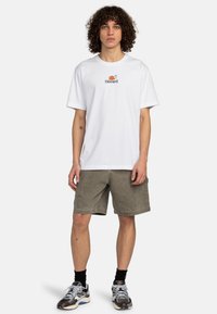 Camiseta blanca de manga corta con el logo "Element" en negro y naranja. Combinada con pantalones cortos verdes oliva y zapatillas negras y grises.