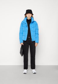 Veste matelassée bleu vif avec un col haut, col roulé noir en dessous, pantalon noir et chapeau seau noir. Tenant un sac texturé noir.