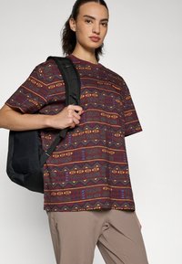 Short-sleeve maroon t-shirt with geometric patterns in yellow, blue, and green. Paired with beige pants and a black backpack.