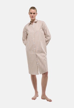 Femme debout pieds nus portant une robe ample, boutonnée, à manches longues et longueur genou avec des rayures verticales beige et marron.