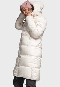 Lange witte pufferjas met verticale stiksels, capuchon en ritssluitingzakken. Gepaard met een lichtroze gebreide muts. Gladde, glanzende textuur.