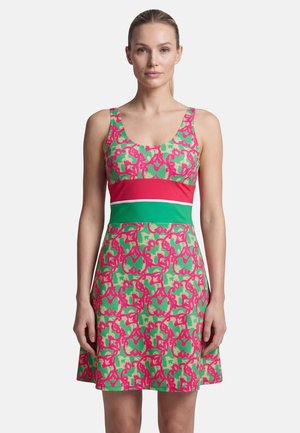 Woman wearing sleeveless dress with green and pink abstract pattern, red and green horizontal chest and waist bands, standing against white background.