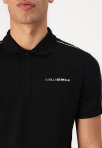Svart polo t-shirt med en treknapplista, guldlogotyp och gula ränder längs axelsömmen. Slät textur och korta ärmar.