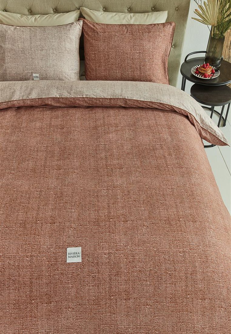 Couvre-lit texturé de couleur rouille avec des oreillers assortis, présentant un motif de tissage subtil. Matériau en lin avec une petite étiquette de marque visible.