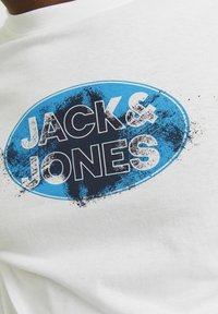 Hvid bomulds t-shirt med en blå oval grafik, der har "JACK & JONES" i fed sort tekst og en slidt struktur omkring kanterne.