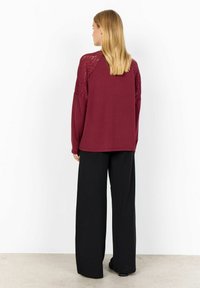 Woman standing facing away wearing a burgundy long-sleeve top with lace shoulders and black wide-leg pants on a plain background.