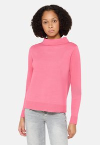 Femme portant un pull à manches longues rose vif et un jean gris clair à pois blancs, debout devant un fond clair uni.