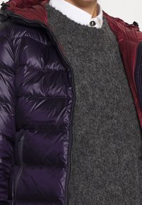 Lichtgewicht donsjack met een kastanjebruine capuchon, een donkerpaarse buitenkant en een grijze gebreide trui eronder. Het jack heeft een met rits afsluitbare zak.