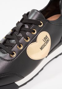 Black leather sneaker with textured heart-shaped logo, gold eyelets, and black laces. Features a white midsole and a smooth finish.