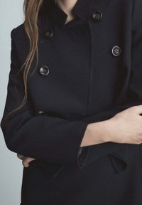 Black double-breasted coat with prominent buttons, structured collar, and tailored fit, featuring smooth fabric and clean lines.