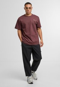 Muž ve hnědém krátkém rukávu, s tmavými volnými kalhotami, bílými teniskami a brýlemi se zlatým rámem, stojící proti jednoduchému světlému pozadí.