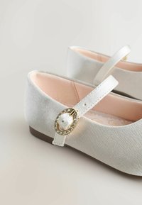 Balerini albe dintr-un material texturat, cu accent de cataramă aurie și căptușeală interioară roz. Curea care se fixează bine în jurul gleznei.