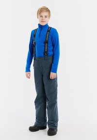 Blauwe fleece pullover met een halflange ritssluiting, gecombineerd met grijze sneeuwbroek met verstelbare zwarte schouderbanden en zwarte waterdichte laarzen.