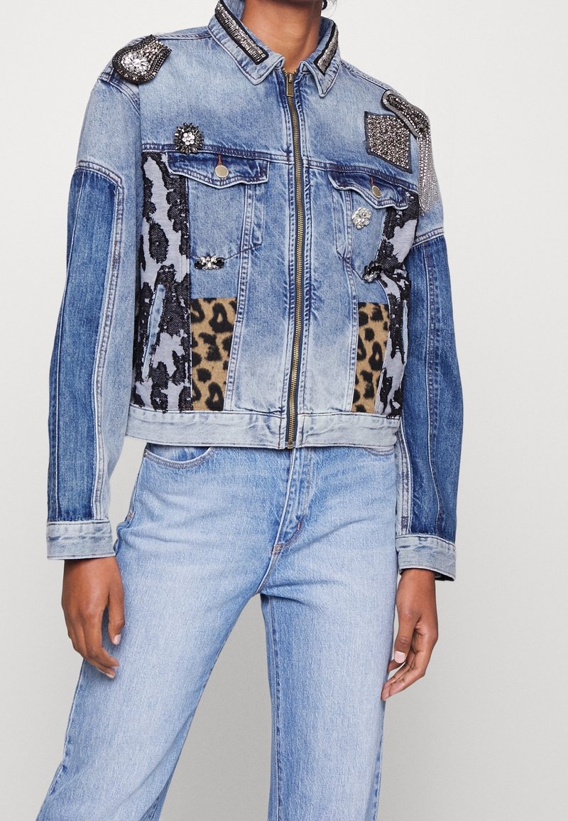 Femme portant une veste en jean bleue avec des patchs léopard et de la dentelle noire, des décorations en strass, et un jean bleu clair sur un fond uni.