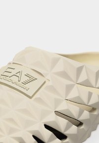 Κοντινό πλάνο σε μπεζ παπούτσι χωρίς κορδόνια με γεωμετρική υφή και πλακέτα με το λογότυπο EA Emporio Armani στο πάνω μέρος.