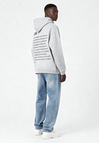 Felpa grigia con testo sul retro, abbinata a jeans chiari strappati e sneakers bianche. La felpa ha un fit rilassato e un cappuccio regolabile.