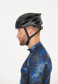 Sort cykelhjælp med aerodynamisk design og flere ventilationsslotte; båret af en person i en blå og sort mønstret trøje.