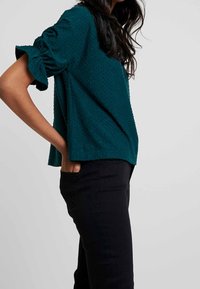 Teal texturerad blus med korta, volangärmar, har en lös passform och rundad nederkant. Kombinationen är med svarta byxor.