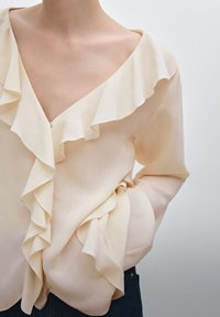 Blusa beige chiaro con scollo a V profondo, maniche lunghe e un design a fronzoli a più strati lungo il décolleté e le maniche.