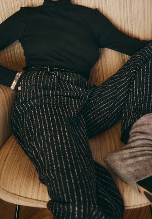 Personne allongée sur une chaise beige rayée, portant un col roulé noir, un pantalon noir à fines rayures, des bottes grises et un bracelet transparent.