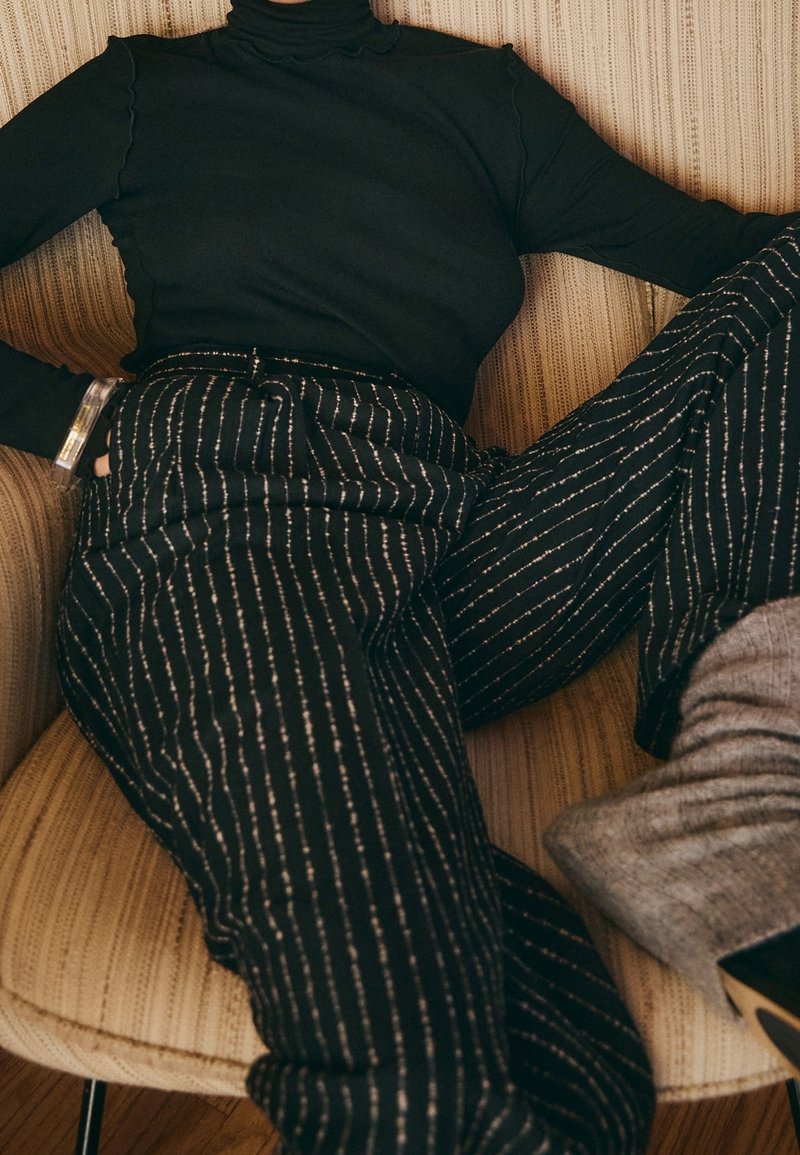 Personne allongée sur une chaise beige rayée, portant un col roulé noir, un pantalon noir à fines rayures, des bottes grises et un bracelet transparent.