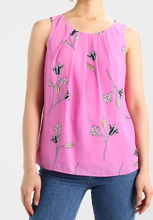 Ærmeløs lyserød bluse med plisseret halsudskæring og sort og beige blomsterprint, båret med blå jeans.