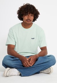 Mann mit lockigem Haar, der mit gekreuzten Beinen sitzt und ein hellgrünes Volcom-T-Shirt, blaue Jeans und weiße Turnschuhe vor einem weißen Hintergrund trägt.