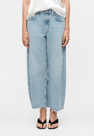 Person iført lyseblå vidde-cropped jeans med rå kanter, hvid top og sorte thongsandaler på hvid baggrund.