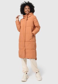Lange oranje pufferjas met capuchon, voorzien van verticale quilting, zijzakken en een ritssluiting. Draag over crèmekleurige broeken en sneakers.