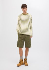 Halenka s dlouhým rukávem se zelenými a krémovými pruhy, v kombinaci s olivovými cargo šortkami. Model má na sobě hnědé boty a šedé ponožky. Minimalistické pozadí.