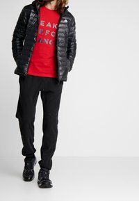 Chaqueta de abrigo negra con capucha, camiseta roja con texto negro y pantalones deportivos negros. Zapatillas negras completan el atuendo.