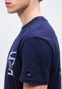 Νεαρό άτομο που φοράει μπλε ναυτικό T-shirt με ένα μικρό κόκκινο, λευκό και μπλε λογότυπο στο μανίκι, εμφανιζόμενο από το πλάι του αυχένα έως το μέσο του βραχίονα.