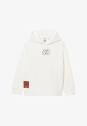 Sweat à capuche blanc Lacoste avec le texte « Roland Garros Club » et un graphisme de filet de tennis centré, ainsi qu'un logo crocodile vert sur un patch marron près de l'ourlet.