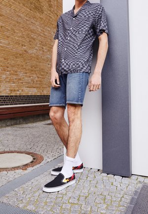 Sort-ærmet sort og grå ternet skjorte, denimshorts, hvide sokker og sorte slip-on sko med flamedesign. Står op ad en væg.