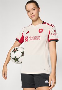 Lys beige fodboldtrøje med røde detaljer, korte ærmer og v-udskæring. Har Liverpool-logoet og teksten "standard Chartered". Sorte shorts.