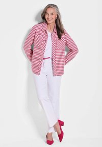Roze en witte geruite jas met kraag, gecombineerd met een wit topje, witte broek en roze hakken; heeft lang haar en een neutrale achtergrond.